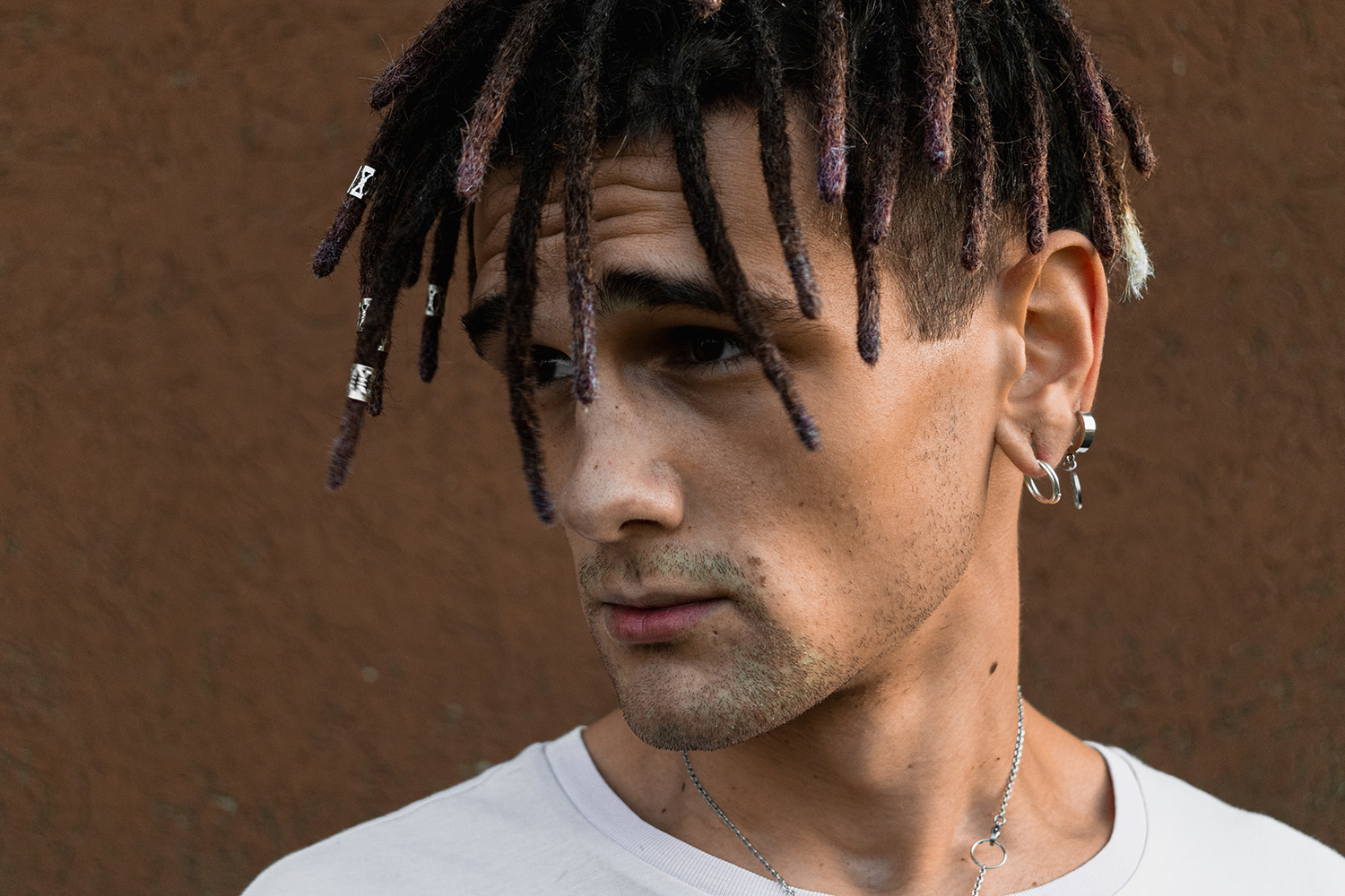 Ritratto di un giovane con dreadlock e orecchini davanti a un muro color terra